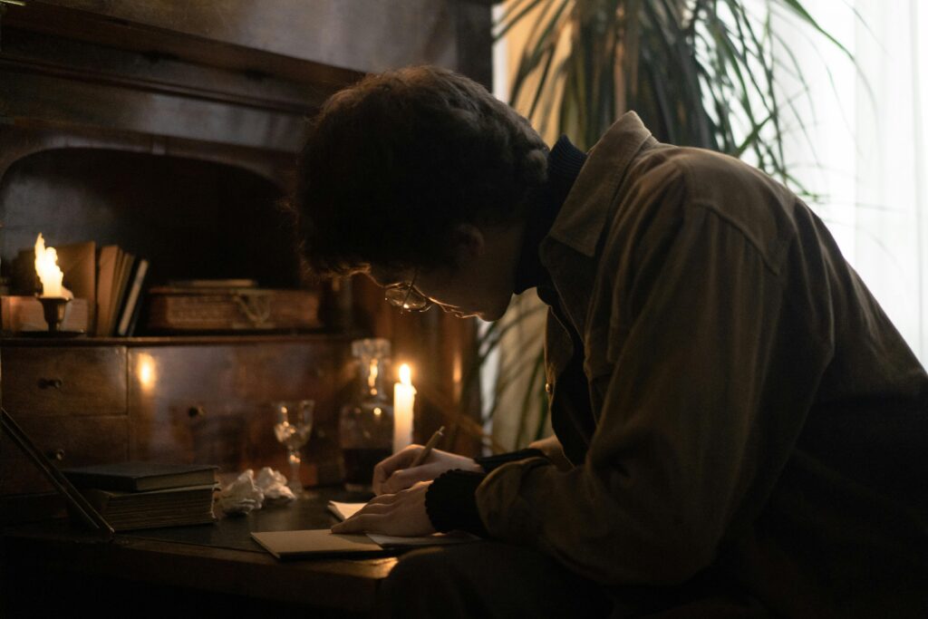 A young man deeply engrossed in writing by candlelight in a cozy study setting.